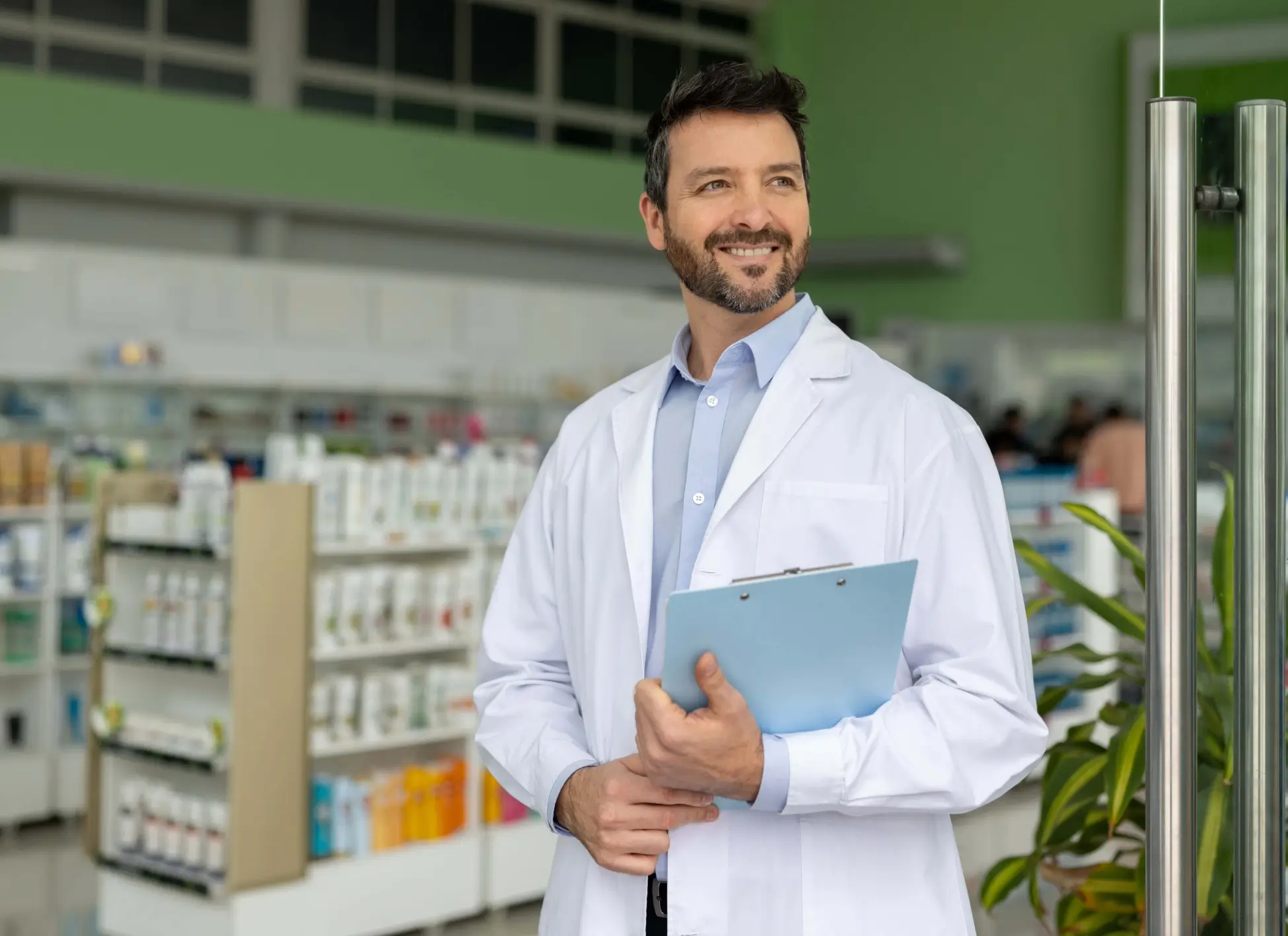A smiling pharmacist in a white lab coat stands in a pharmacy, holding a clipboard. In the background are shelves of medicine