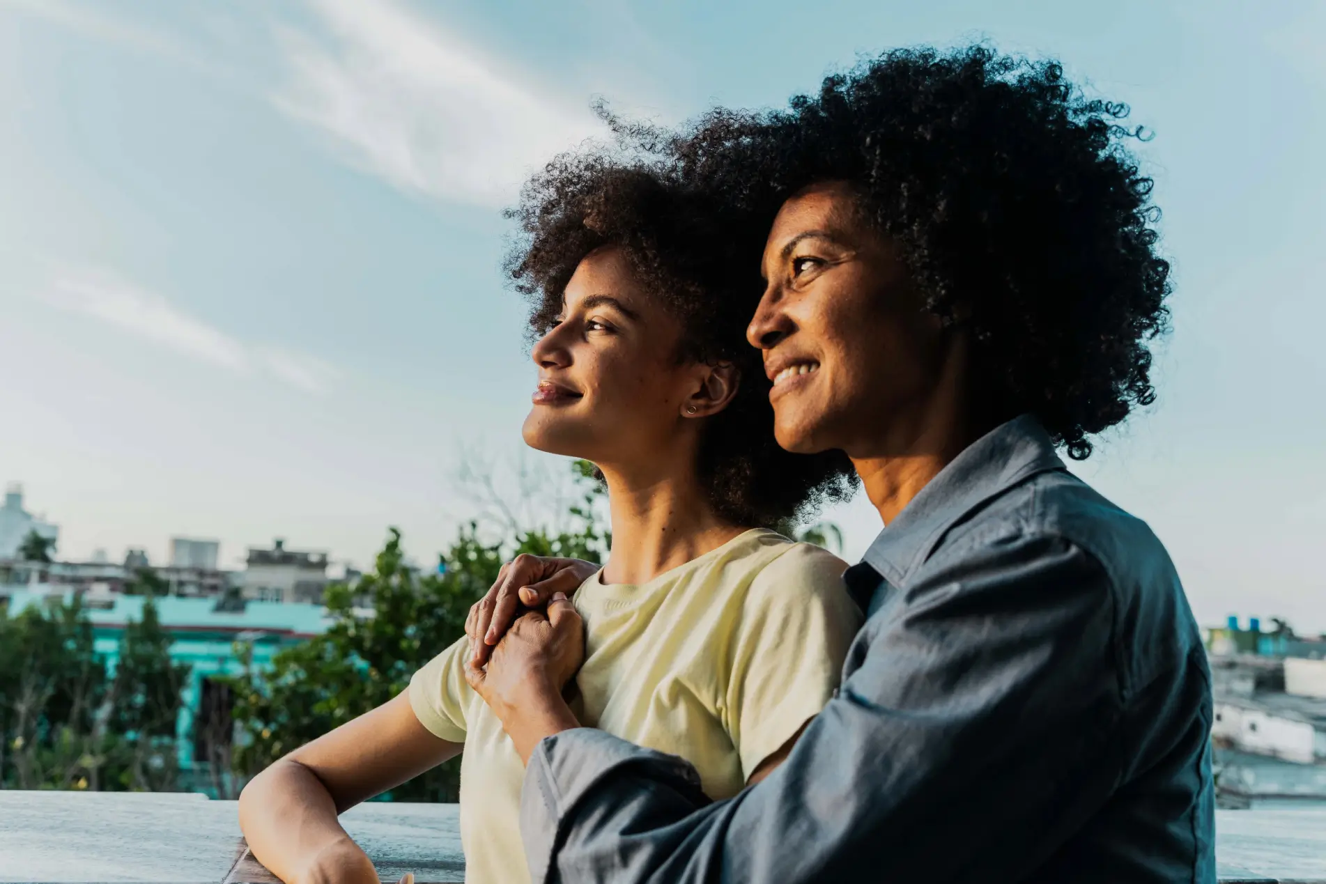 A woman with an arm around a younger woman, both smiling and looking into the distance with a cityscape in the background.