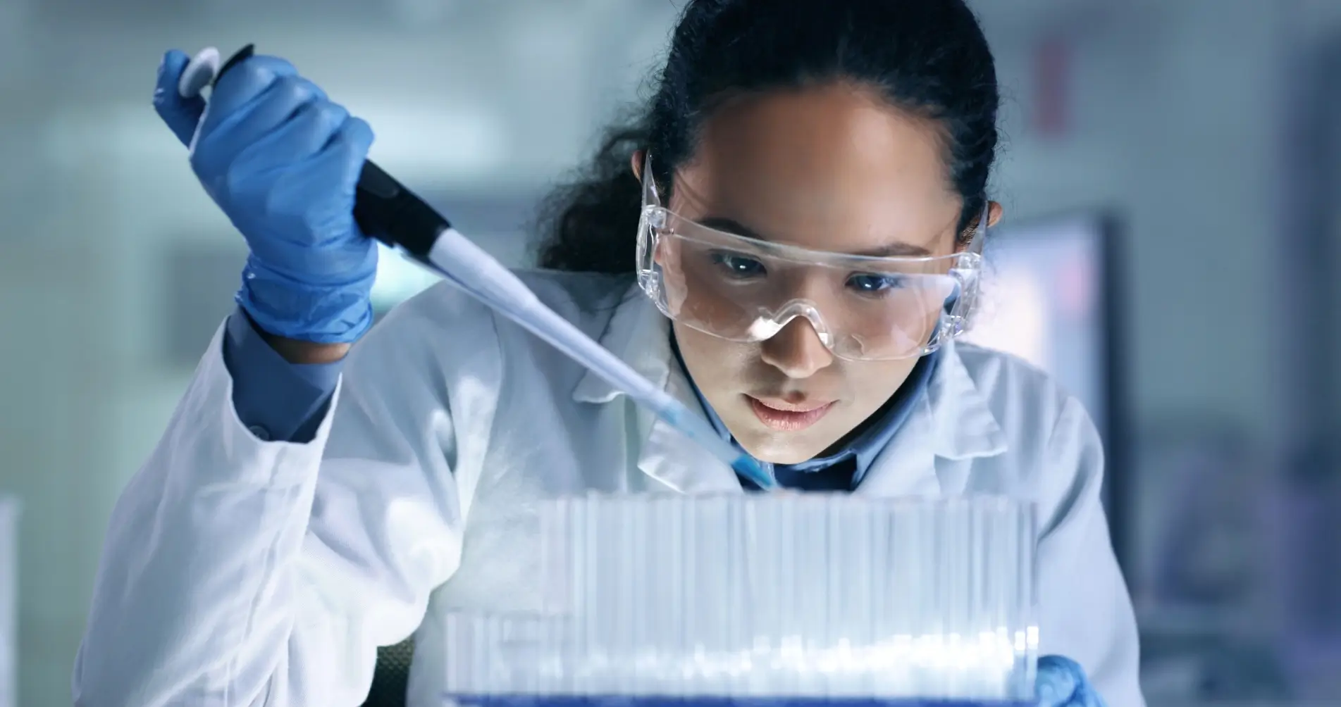 A scientist wearing goggles and gloves uses a pipette in a laboratory setting.