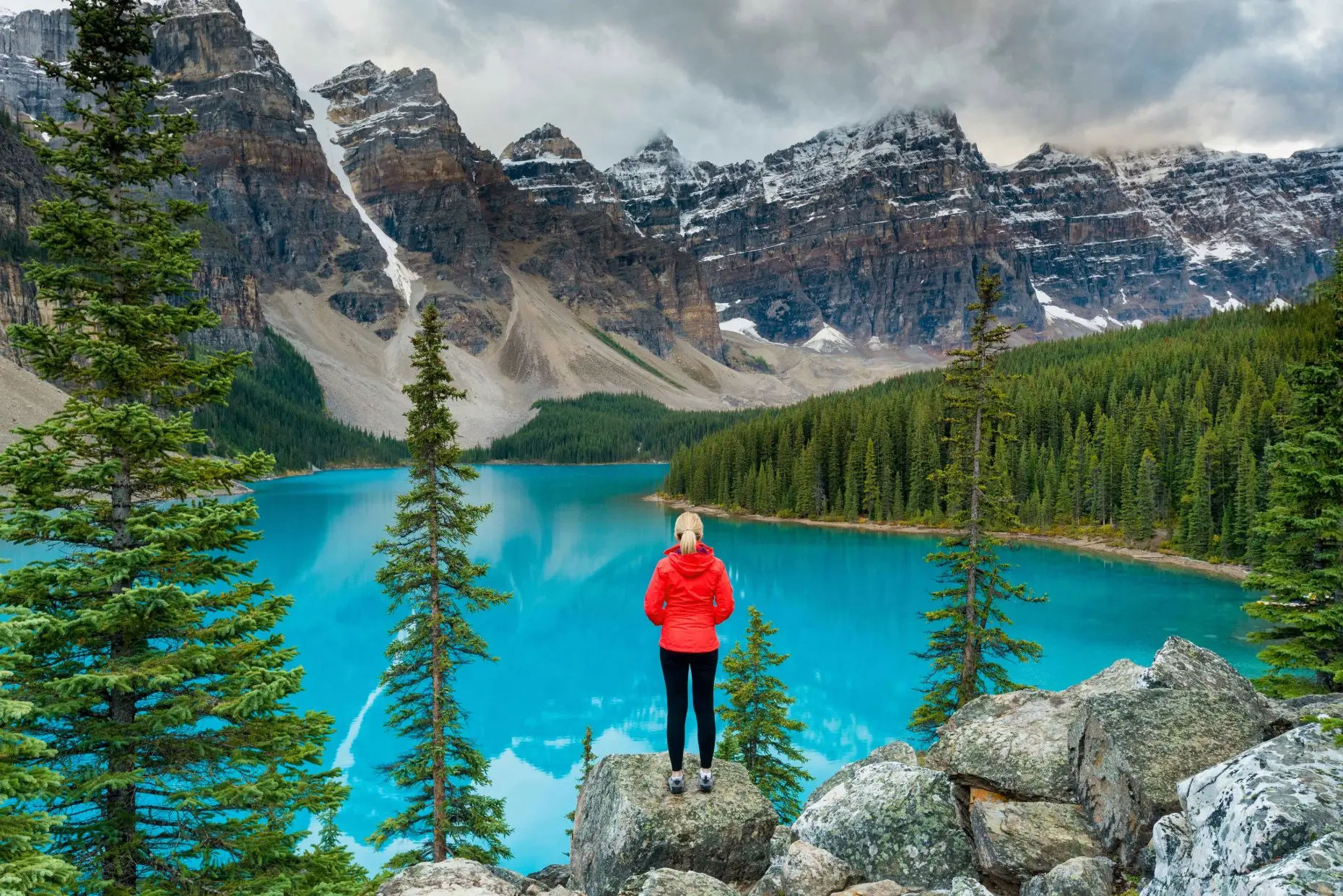 A person in a red jacket stands on rocks overlooking a turquoise lake surrounded by evergreen trees and snow-capped mountains under a cloudy sky.
